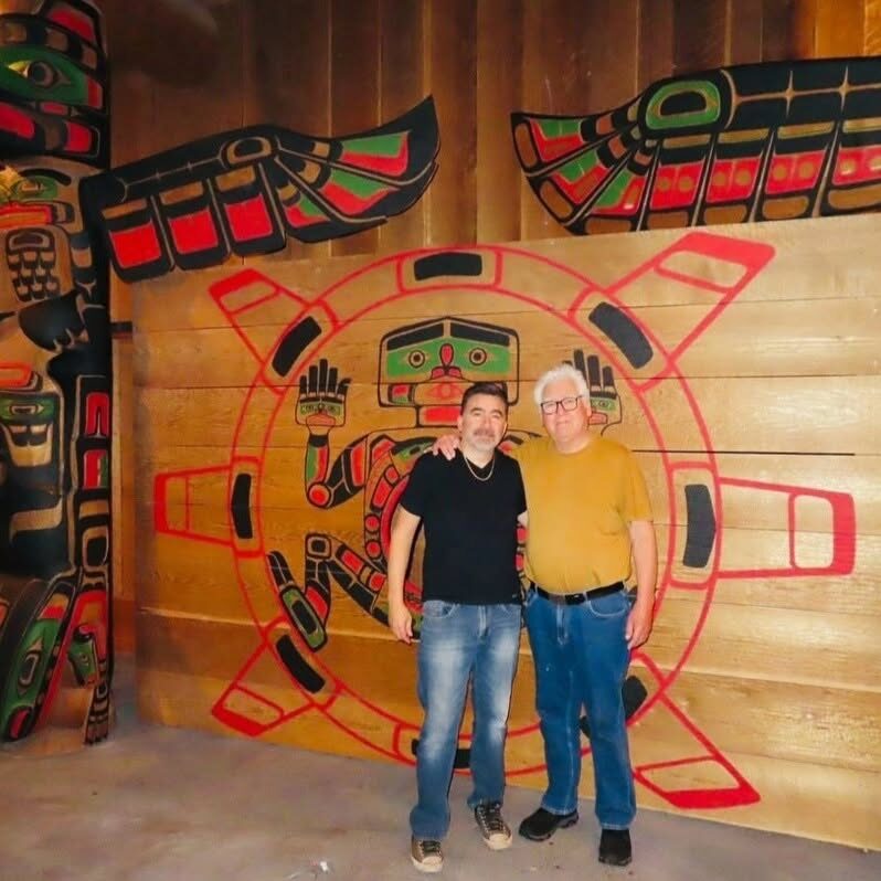 Two men in front of a large red, black and green painted screen inside a cedar bighouse, with a totem pole at left