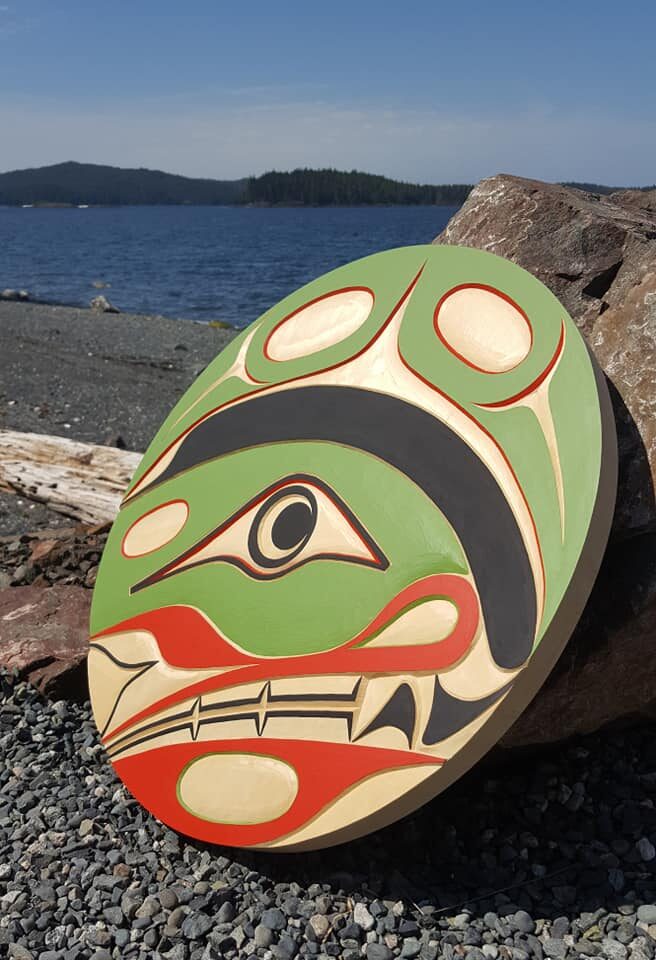 Round panel painted in green, red, black and cream, leaning on driftwood and rock at a pebble beach by the ocean