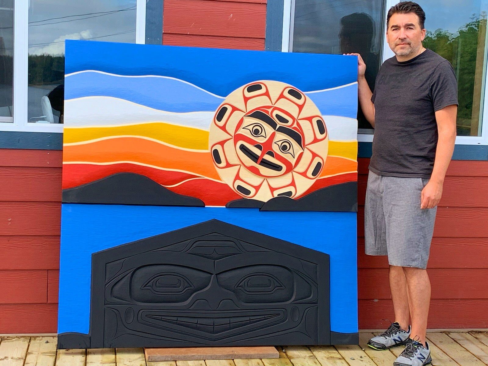 Jason standing beside a painted sun-and-waves panel above a black carved house-front design, propped against a red wall
