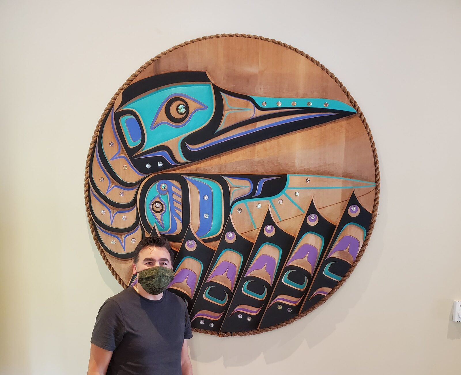 Jason beside a large round hummingbird panel carved in cedar and painted in turquoise, blue and purple, edged with rope