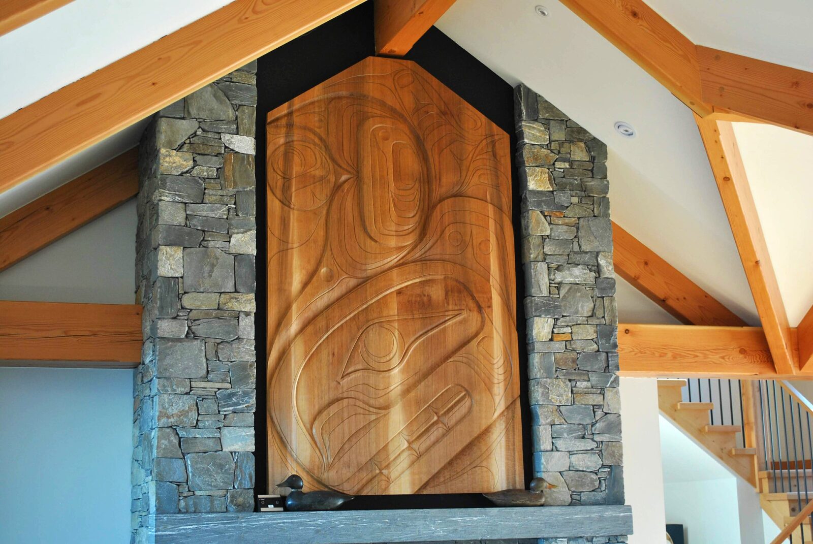 Tall unpainted cedar panel with deep-carved formline set into a stone chimney beneath timber beams